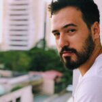 A serious-looking man with a beard stands on a balcony with blurred city buildings in the background.